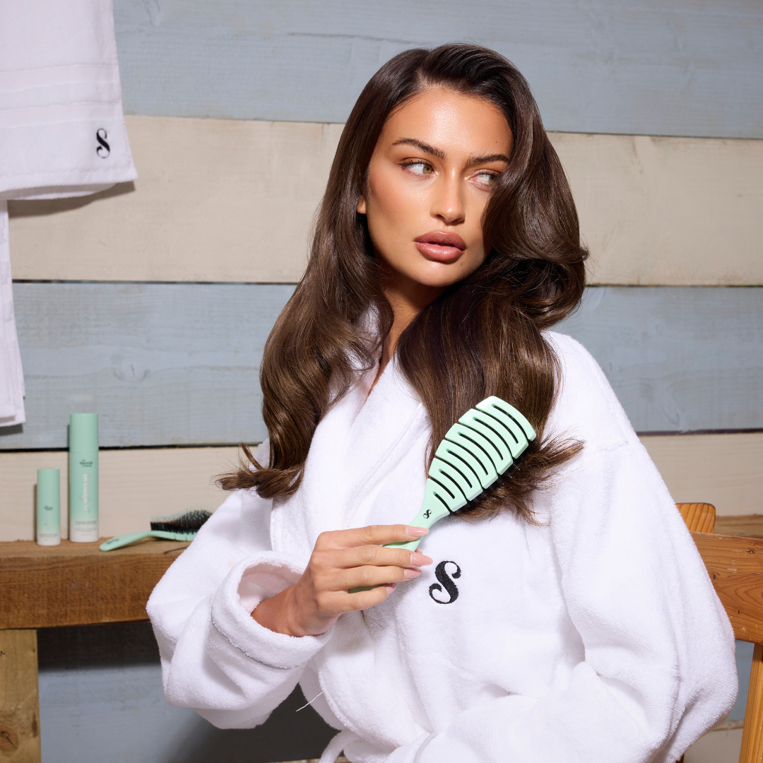 Woman in a white robe brushing her hair with a teal brush in a bathroom setting.