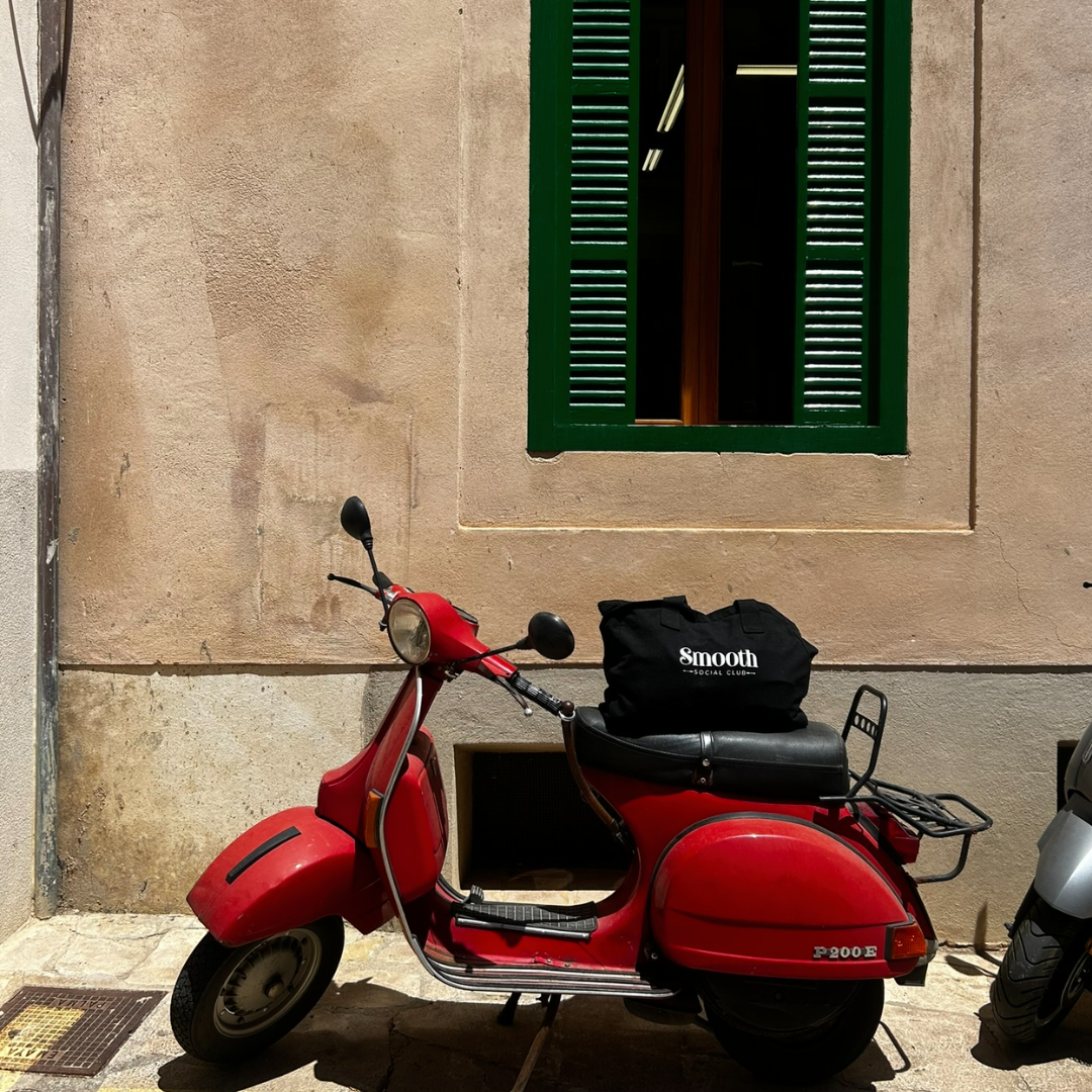 Red scooter with a black bag on a sidewalk next to a building with green shutters. Smooth Tote bag is placed on top of the scooter. 