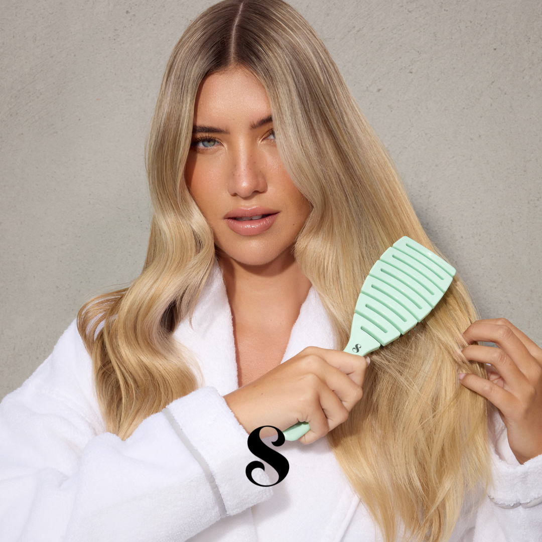 Woman brushing her blonde hair with a teal brush against a neutral background